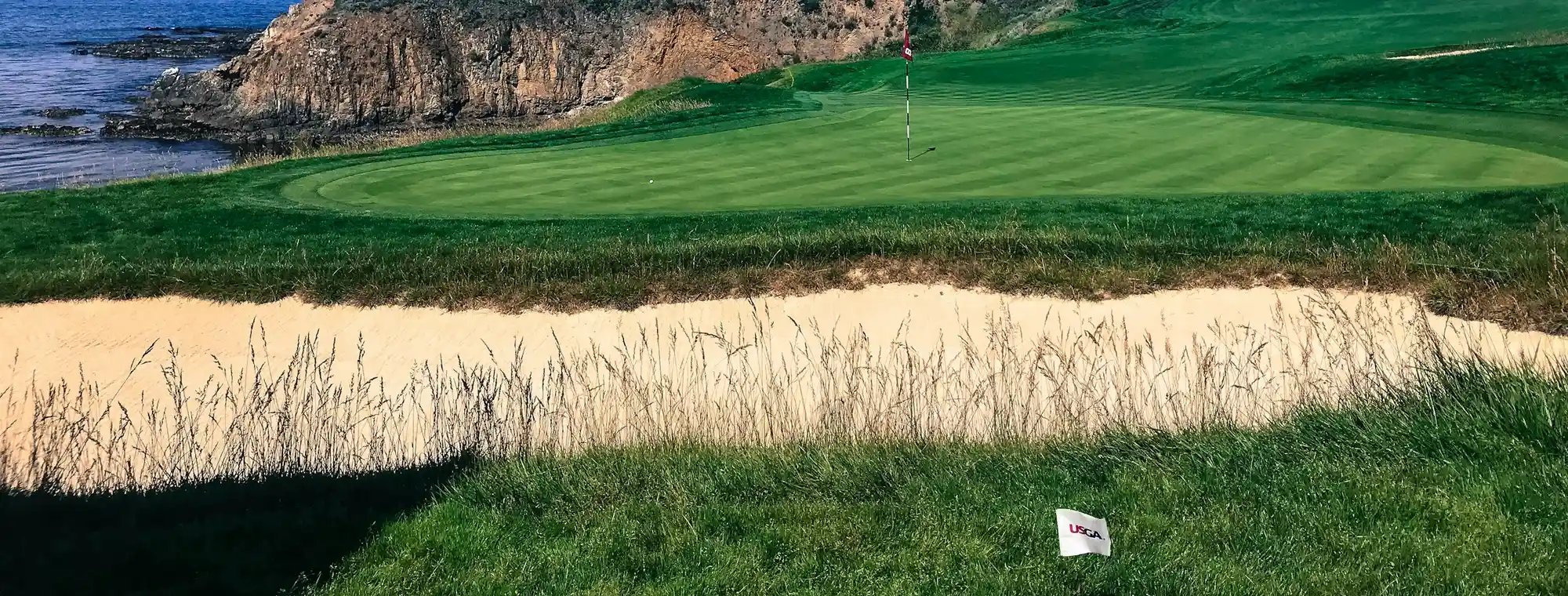 A clifftop links green with a pin and sand bunker perched above a rugged rocky coastline and ocean.
