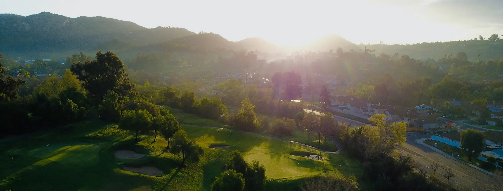 An aerial view of a lush golf-course green and bunkers with the low sun flaring over the hills at golden hour.
