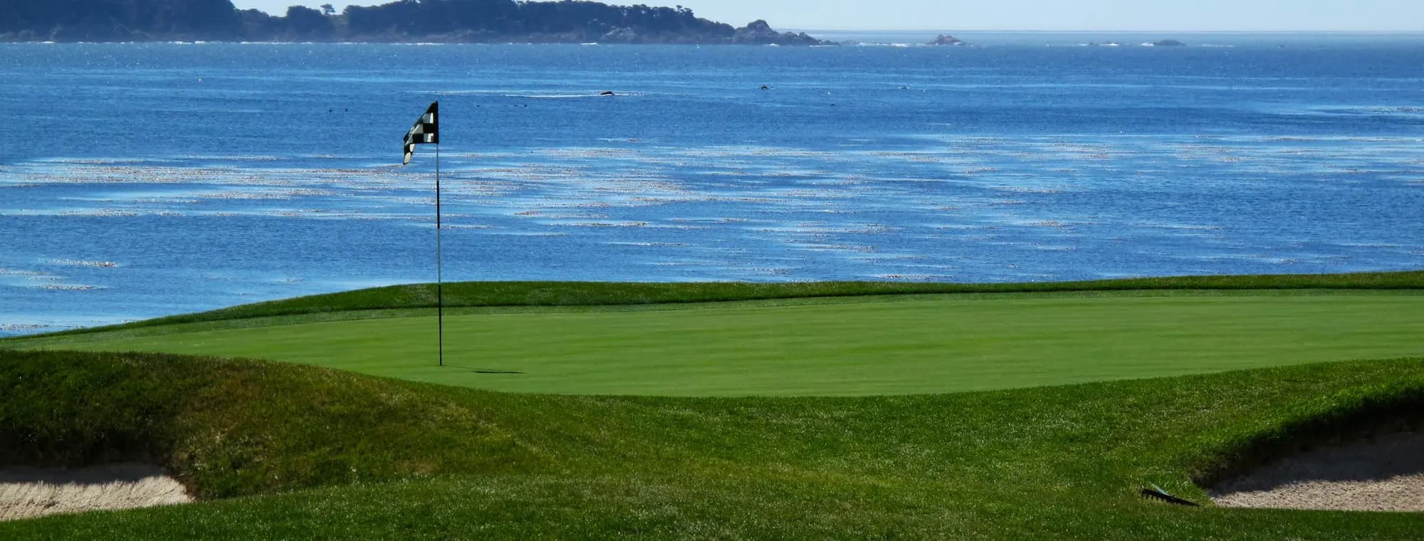 A scenic par-3 golf hole with a green jutting out over calm blue water, framed by trees.