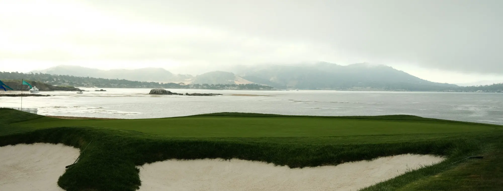 An elevated coastal golf-course vista looking over a green and bunkers out to the bay and misty hills.